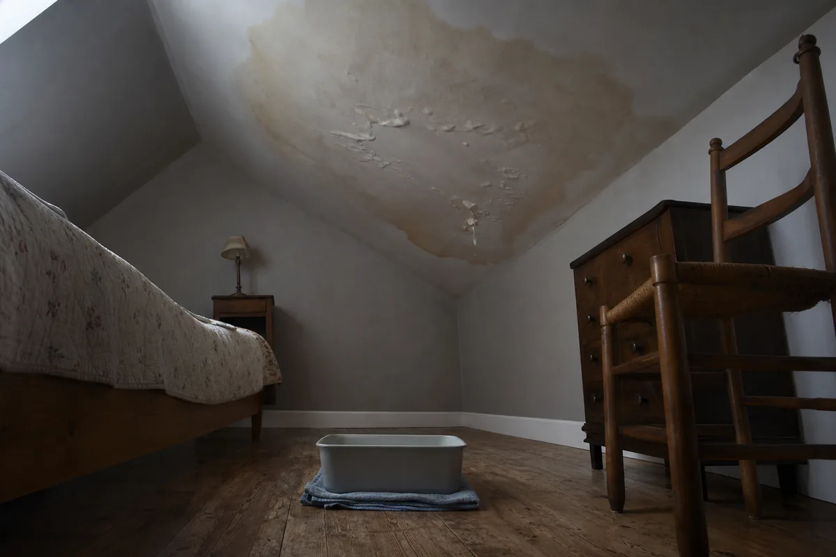 Auréole d’infiltration brune sur le plafond d’une chambre sous combles avec un bac posé au sol