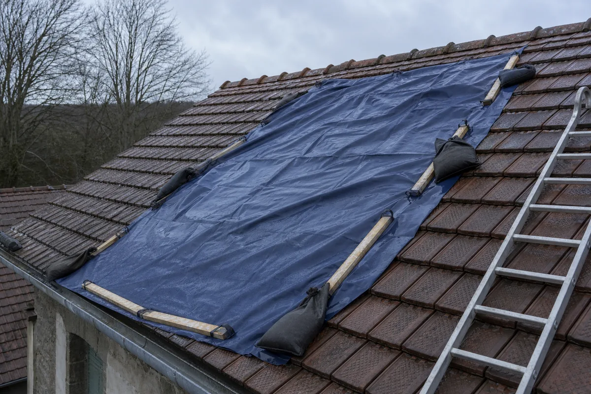 Bâche de protection lestée et sanglée sur un versant de toiture en tuile après une infiltration